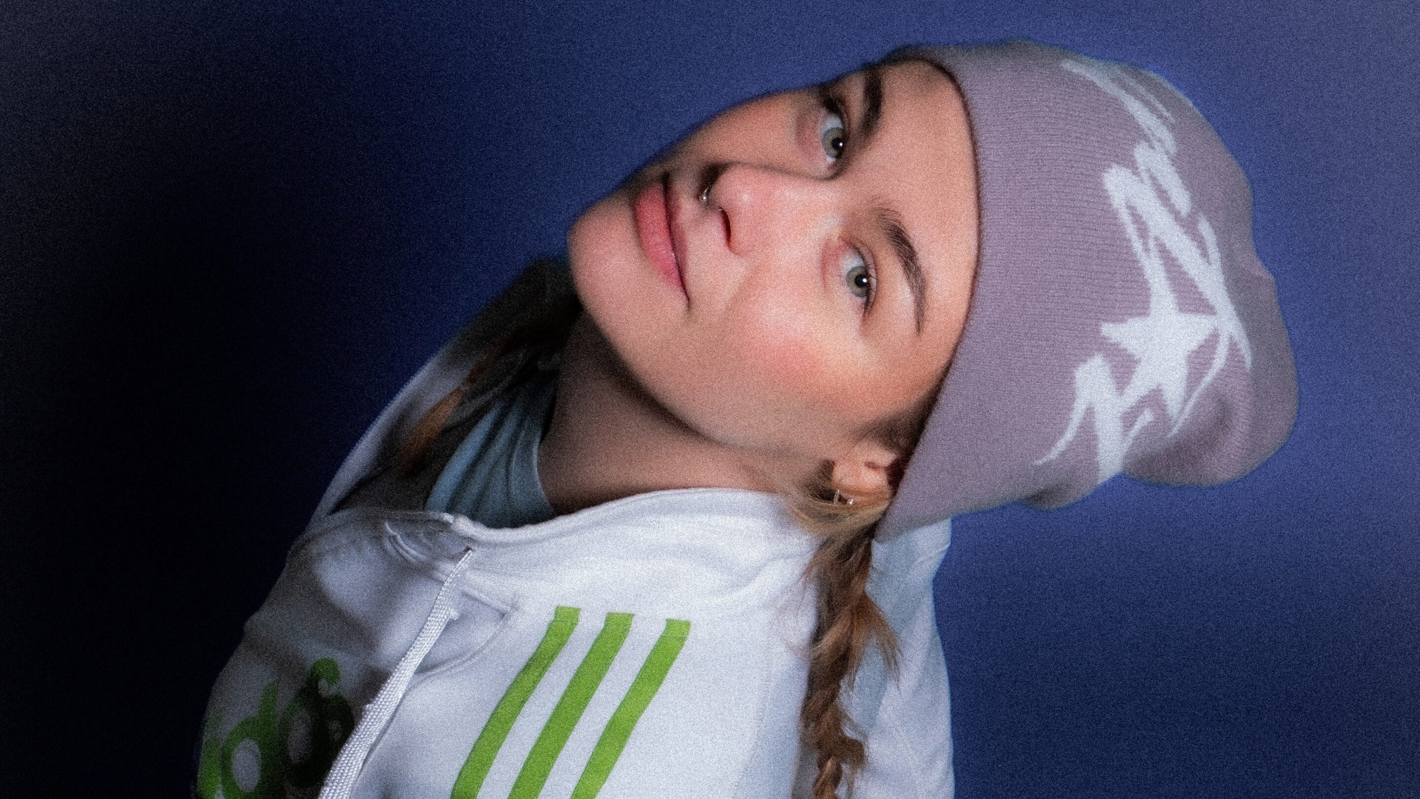 Kvinde i hvid hættetrøje med grønne striber og lyslilla hue, der lægger hovedet på skrå og kigger ind i kameraet mod en ensfarvet lilla baggrund.