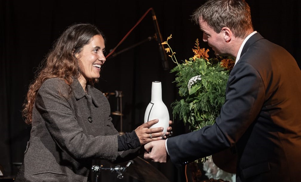 En kvinde smiler, da hun modtager en hvid vaseformet pris fra en mand i jakkesæt på scenen med en buket blomster i baggrunden.