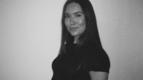 En kvinde med langt, glat hår og et lille smil står op ad en almindelig væg iført en mørk, kortærmet top. Billedet er i sort-hvid.