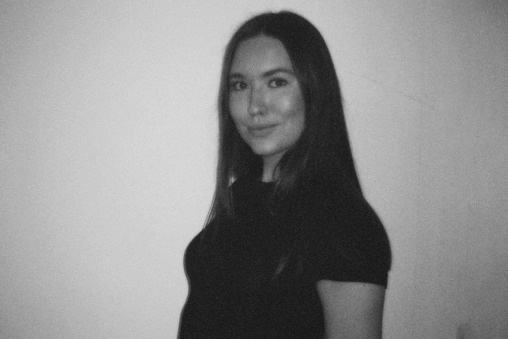 En kvinde med langt, glat hår og et lille smil står op ad en almindelig væg iført en mørk, kortærmet top. Billedet er i sort-hvid.