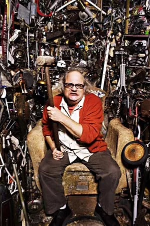 En mand med briller og rød cardigan sidder i en gammel lænestol med en forhammer i hånden, omgivet af et stort udvalg af forskelligt mekanisk skrammel.