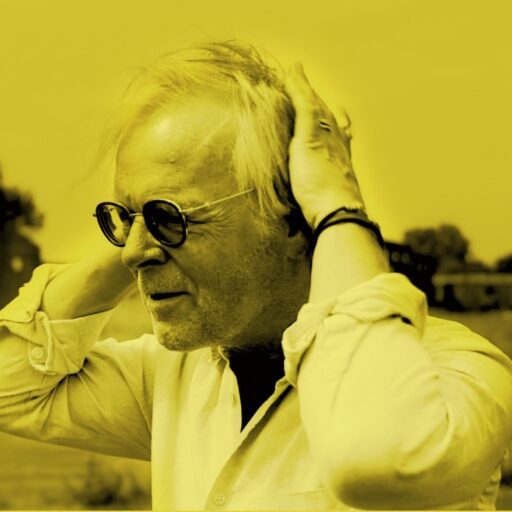 En ældre mand iført solbriller og en lys skjorte står udendørs og holder sit hoved med begge hænder. Billedet har et gult skær.