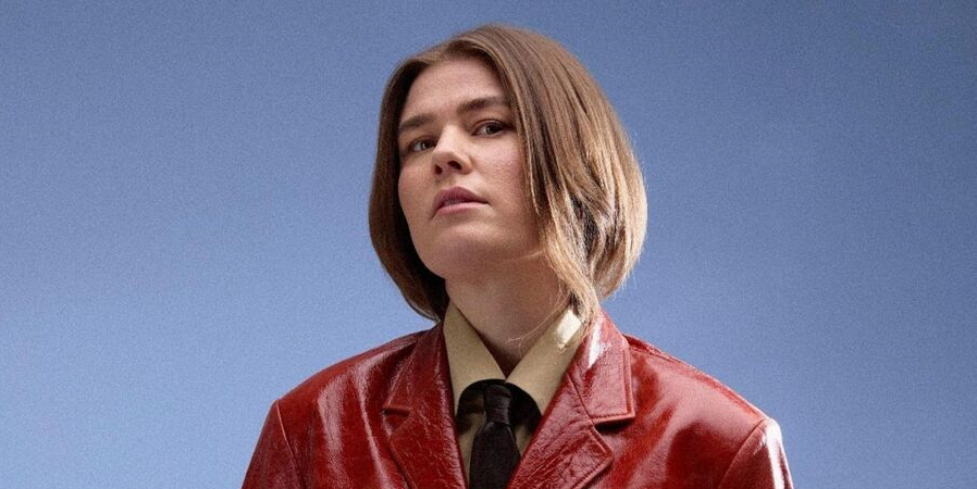 En person med glat, hagelangt brunt hår bærer en skinnende rød jakke over en solbrun skjorte og et mørkt slips og står på en almindelig blå baggrund.