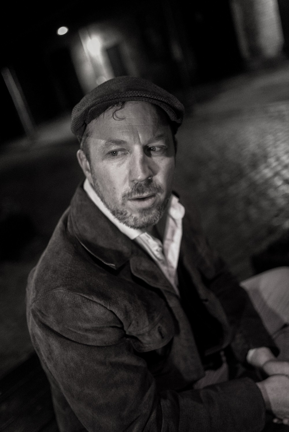 En mand iført flad kasket og jakke sidder udendørs om natten og kigger til siden med et alvorligt udtryk; billedet er i sort-hvid.