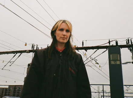 En person med langt brunt hår og lyse striber står udendørs foran elektriske togledninger og master under en overskyet himmel.