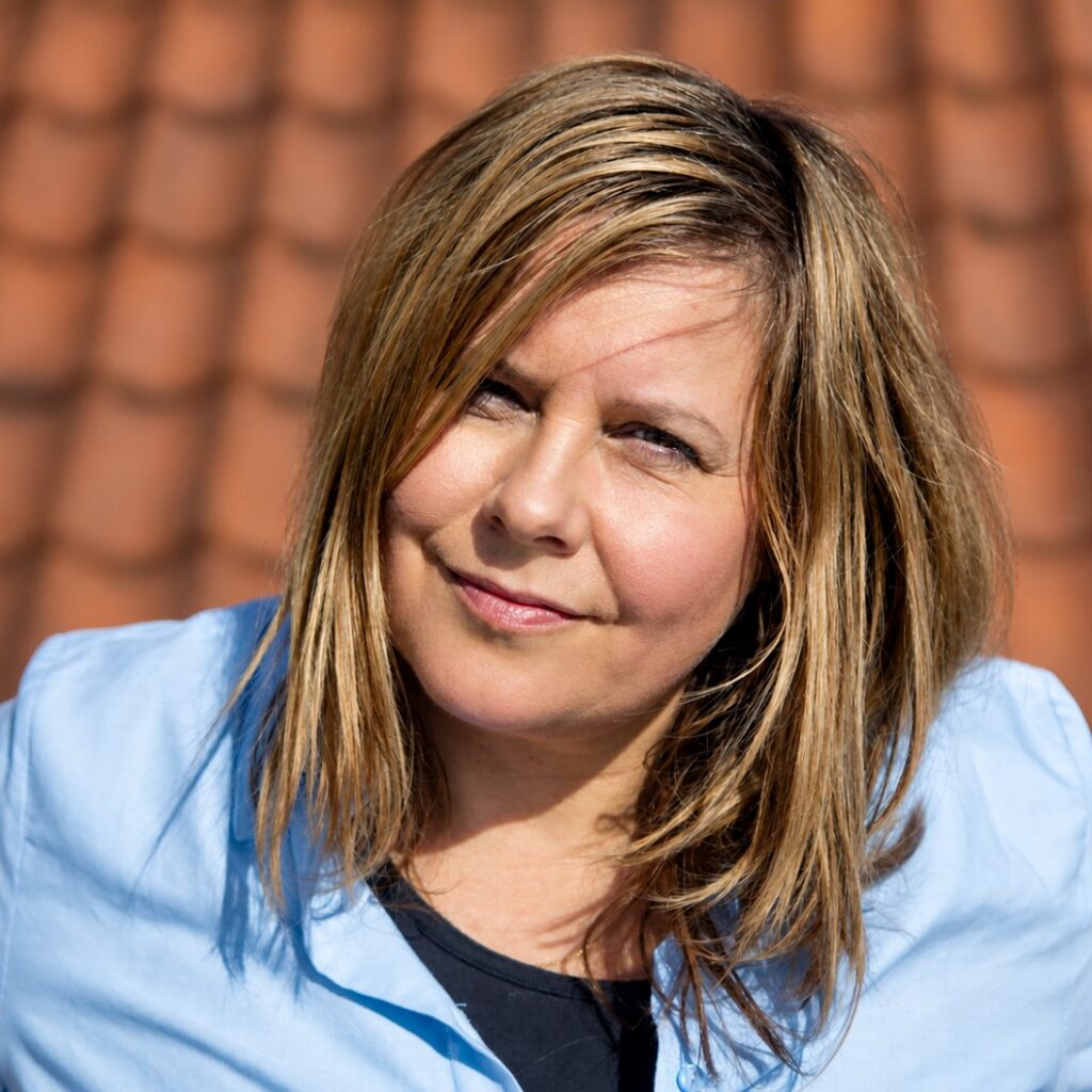 En kvinde med skulderlangt blondt hår, iført en lyseblå skjorte, står foran en baggrund af røde tagsten og kigger ind i kameraet.