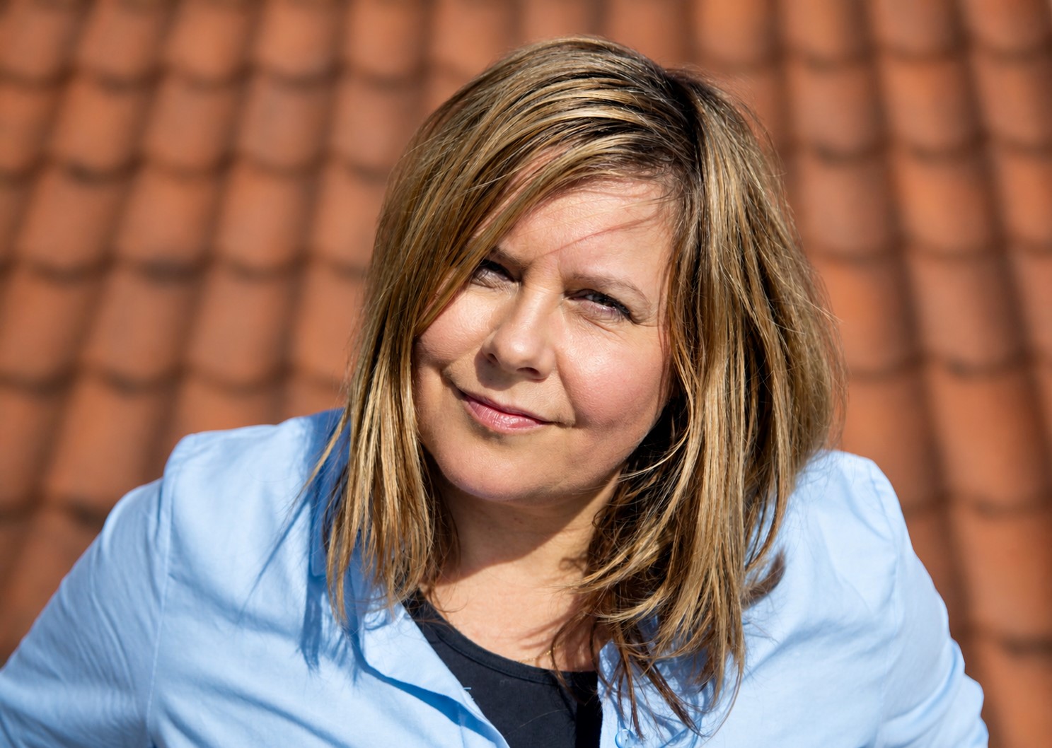 En kvinde med skulderlangt blondt hår, iført en lyseblå skjorte, står foran en baggrund af røde tagsten og kigger ind i kameraet.