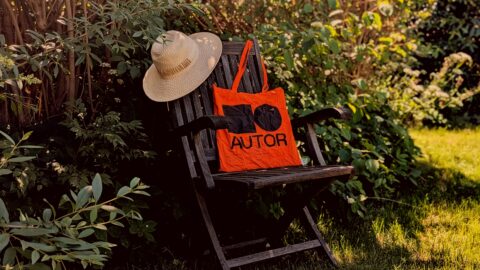 En stråhat og en orange indkøbspose med ordet »AUTOR« ligger på en træstol i en solbeskinnet have omgivet af grønne planter.