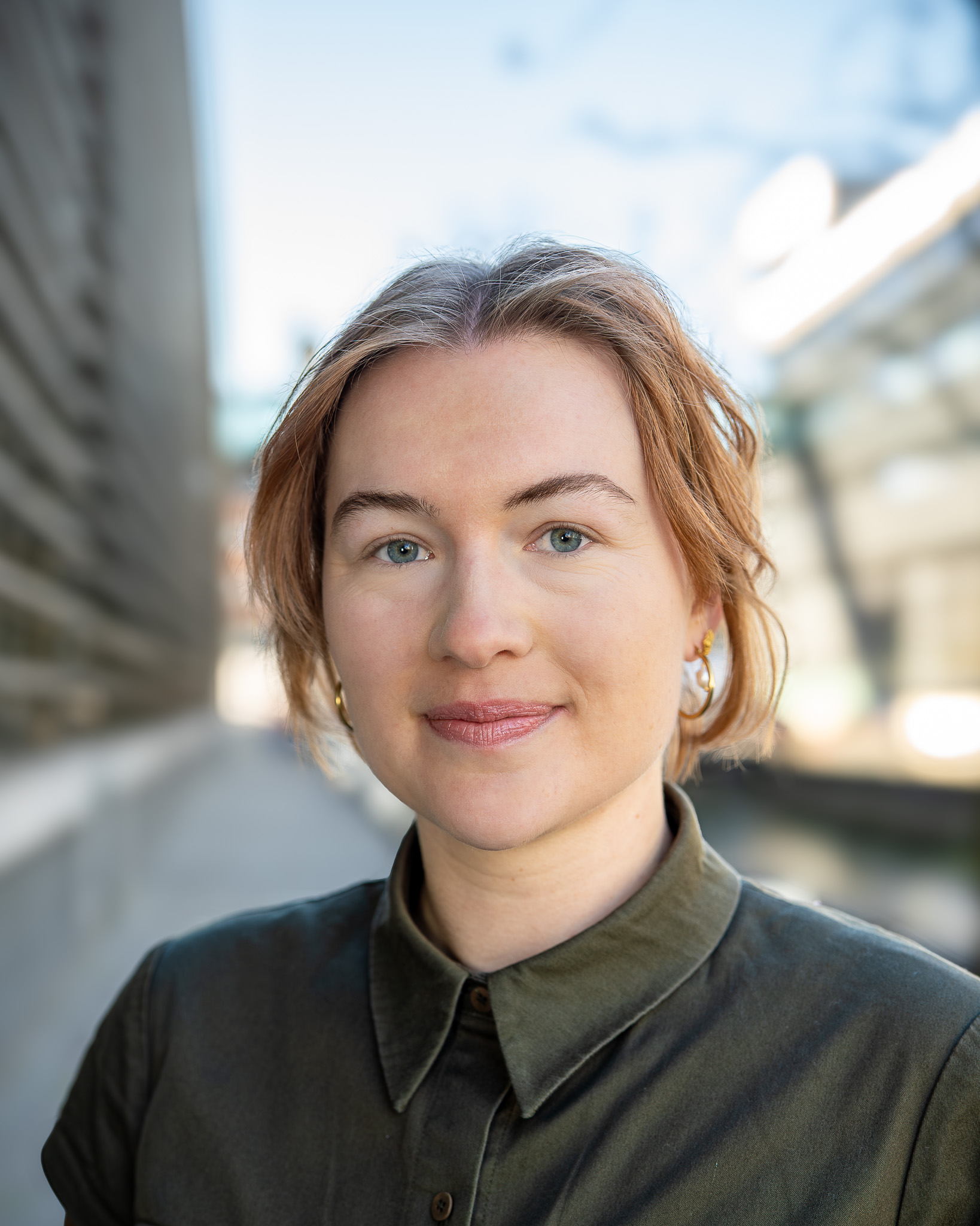 En person med kort, lysebrunt hår og grønne øjne, iført en mørk skjorte med krave, står udendørs med moderne bygninger i baggrunden.