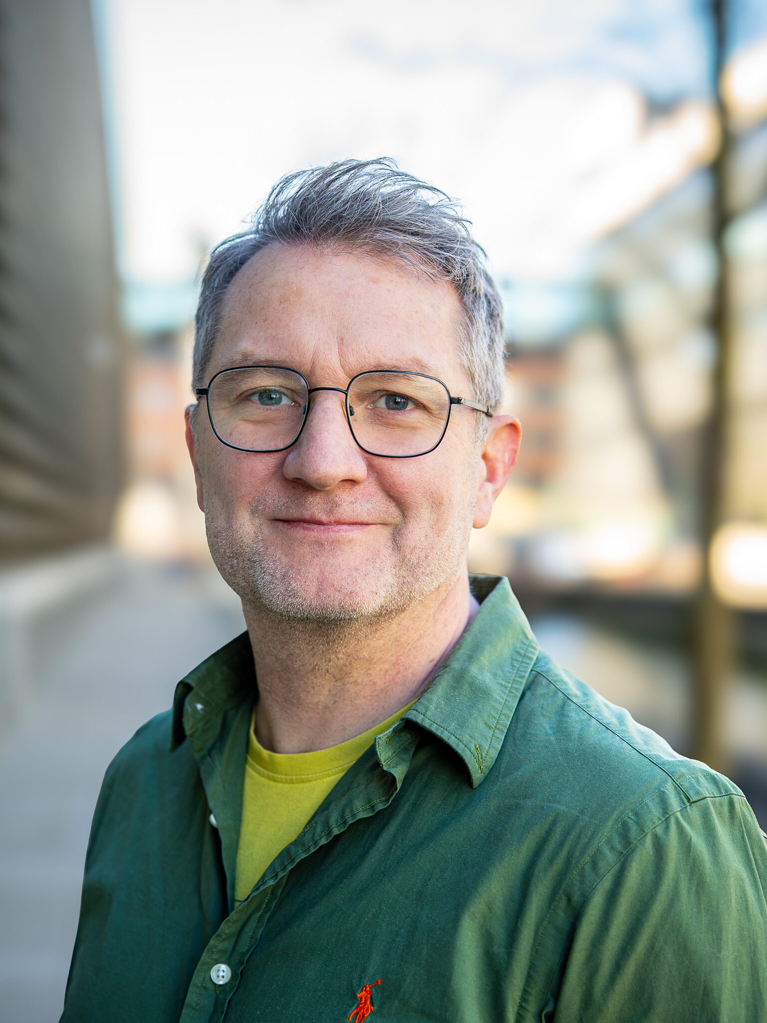 En mand i middelalderen med gråt hår og briller, iført en grøn skjorte, står udendørs med slørede bygninger i baggrunden.