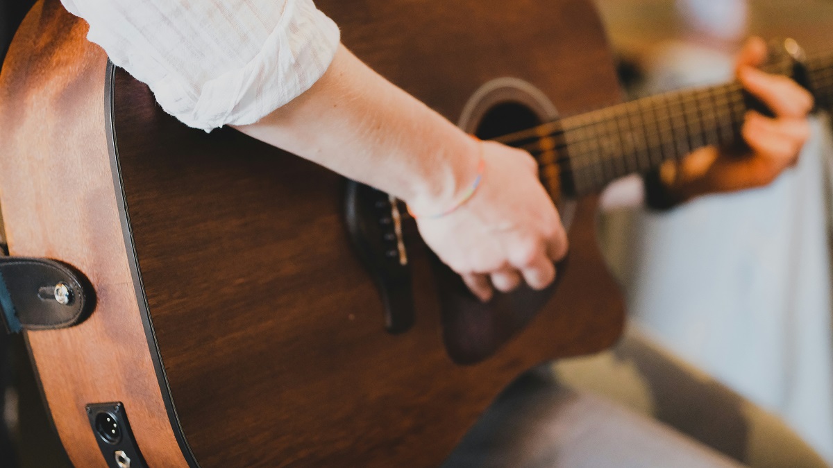 En person iført en hvid skjorte spiller på en akustisk guitar, og fokus er rettet mod den hånd, der strummer på strengene.