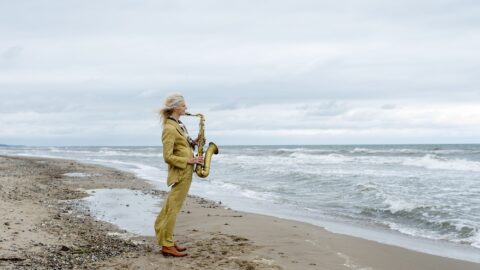 En person i et gult jakkesæt står på en sandstrand og spiller saxofon, mens han kigger ud mod havet under en overskyet himmel.