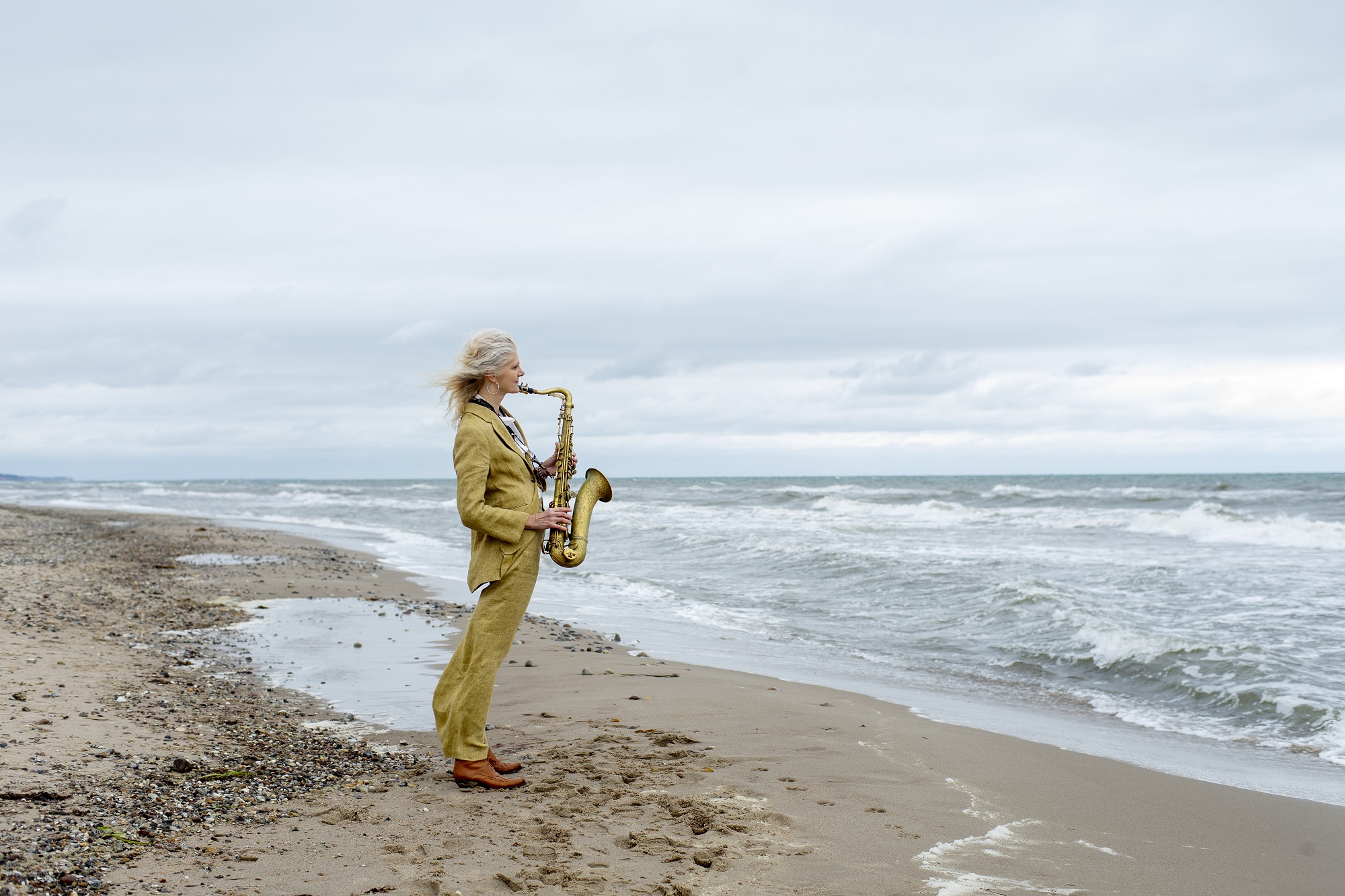 En person i et gult jakkesæt står på en sandstrand og spiller saxofon, mens han kigger ud mod havet under en overskyet himmel.