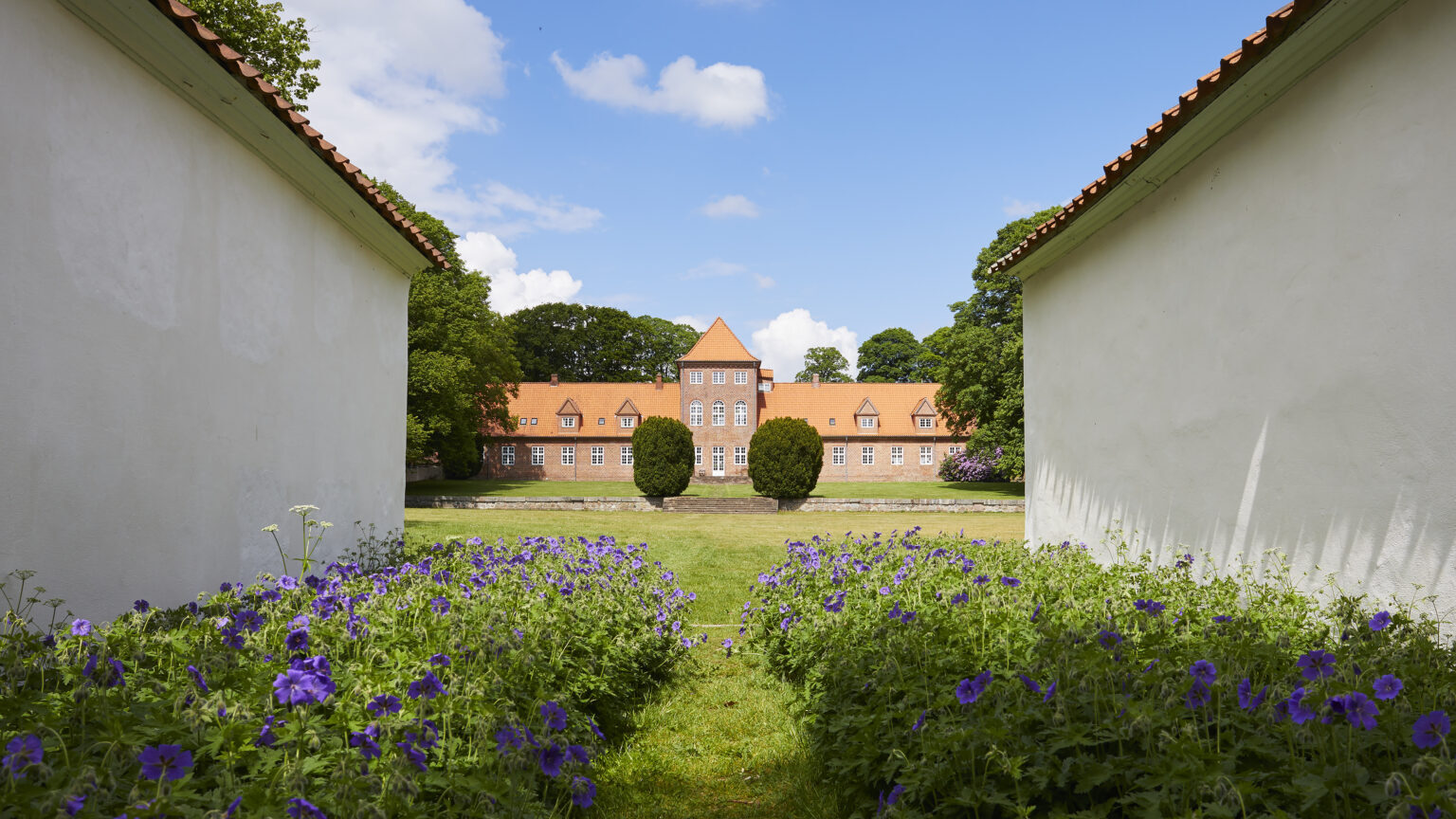 Udsigt mellem to hvide bygninger med en sti omkranset af lilla blomster, der fører til en stor murstensbygning med rødt tag i baggrunden under en blå himmel med skyer.