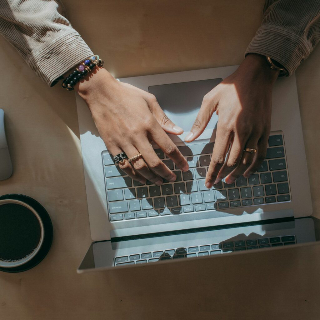 En person, der sidder ved et skrivebord og skriver på en bærbar computer, med en kop kaffe, en trådløs mus og en stak farverige mapper.