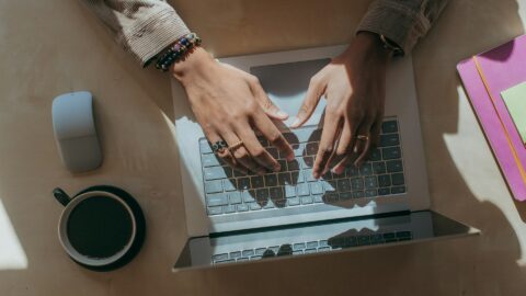 En person, der sidder ved et skrivebord og skriver på en bærbar computer, med en kop kaffe, en trådløs mus og en stak farverige mapper.