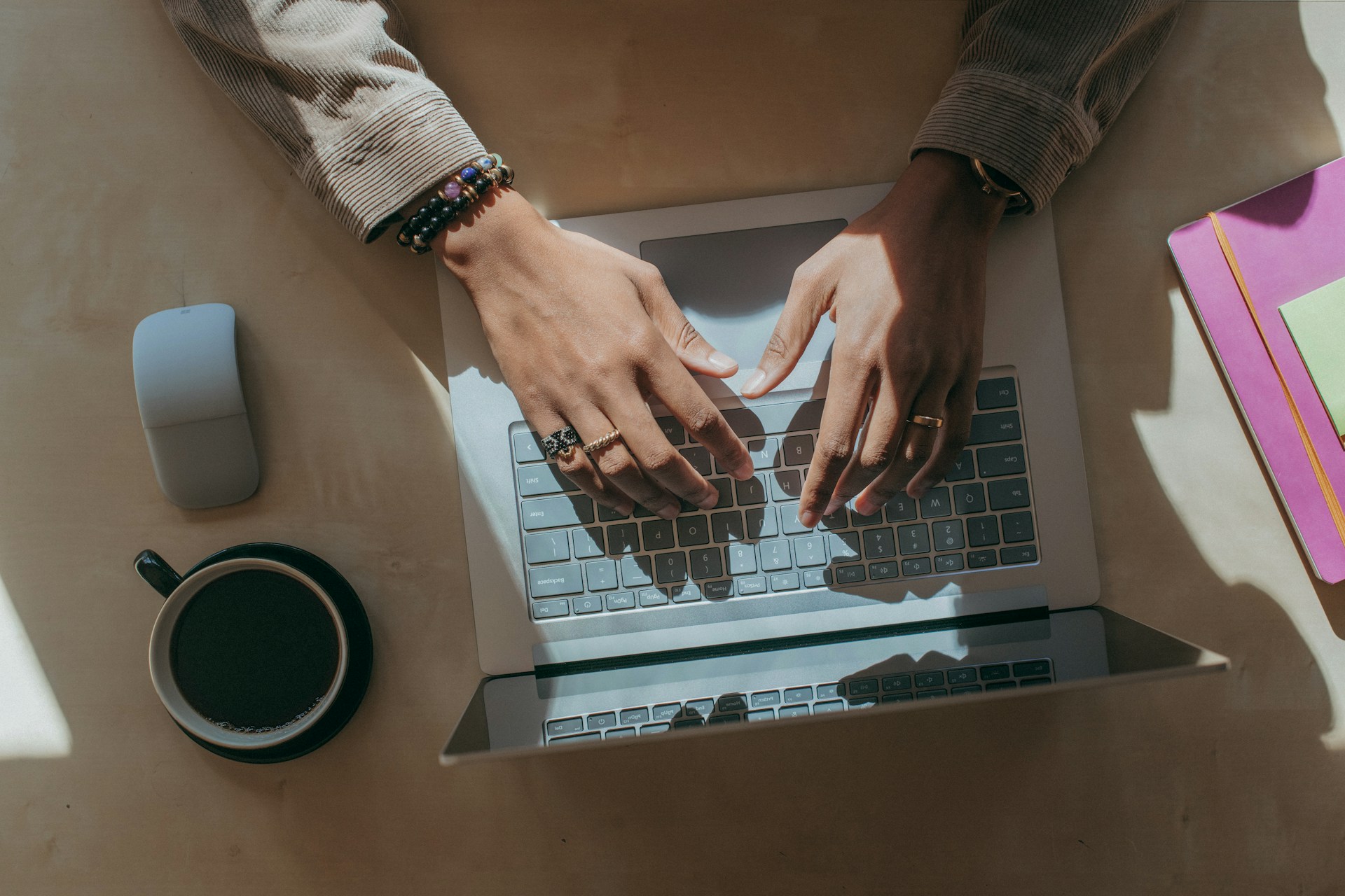 En person, der sidder ved et skrivebord og skriver på en bærbar computer, med en kop kaffe, en trådløs mus og en stak farverige mapper.