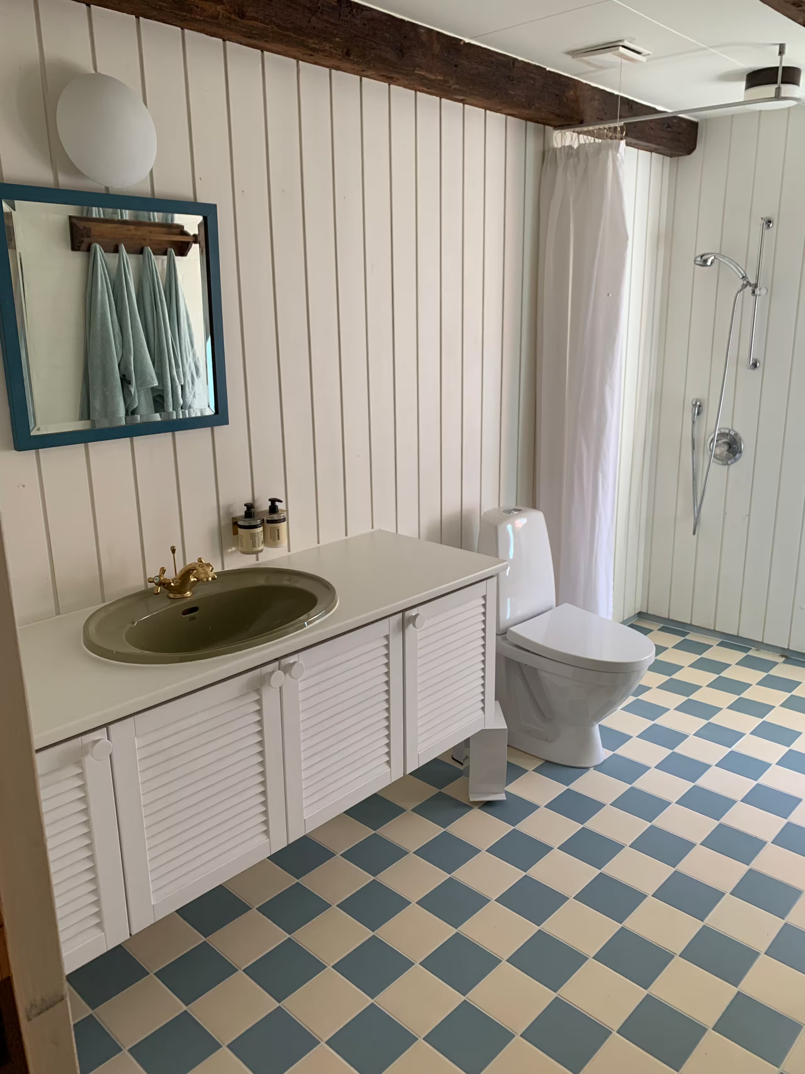 Badeværelse med hvide paneler på væggene, et gulv i blå og hvide tern, en grøn håndvask på et hvidt badeværelsesmøbel, et toilet og en brusekabine med et forhæng og to håndklæder hængende på væggen.