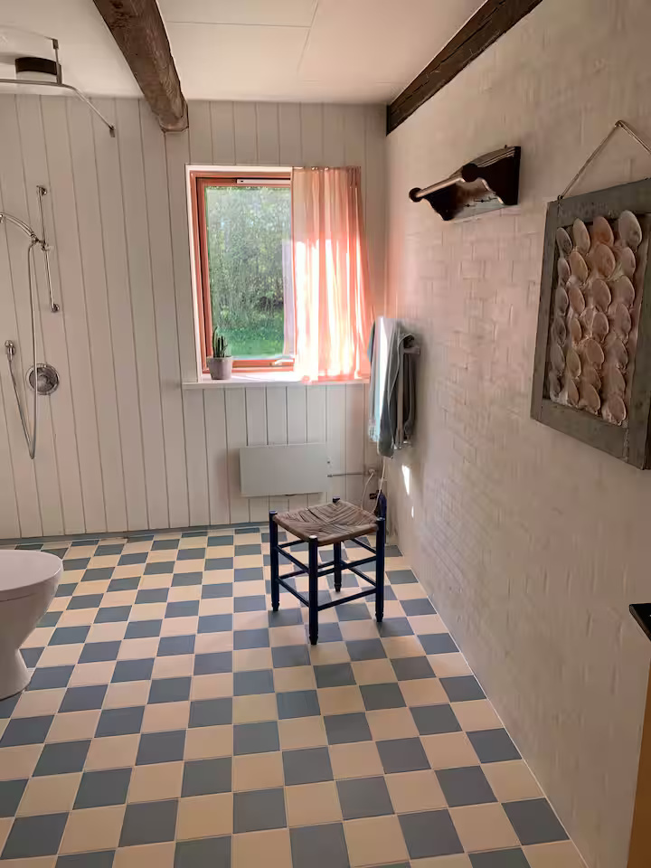 Badeværelse med hvide vægge, et gulv med blå og hvide fliser i tern, et vindue med lyserøde gardiner, en stol, et håndklæde, dekorationer af muslingeskaller og et brusebad.