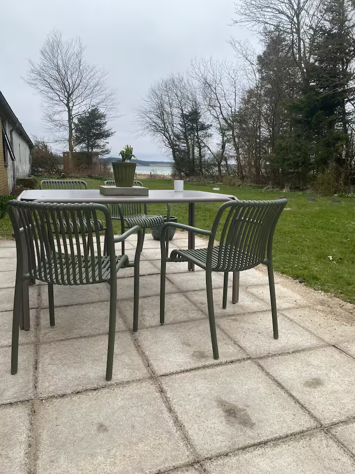 Fire grønne metalstole og et bord med en potteplante på en terrasse med udsigt over en græsplæne og vandet i det fjerne, med træer i baggrunden.