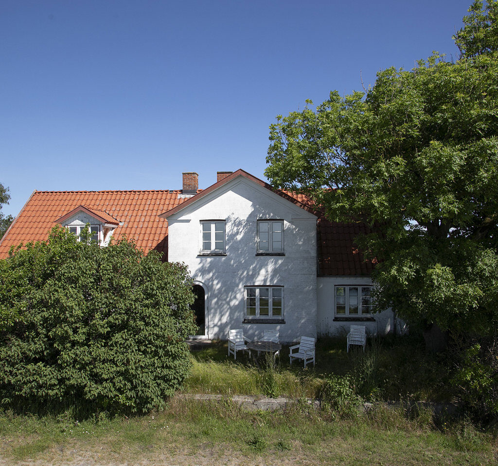 Et hvidt hus med rødt tegltag, omgivet af grønne buske og træer, står under en klar blå himmel. Tæt ved huset ses en hvid stol og et bord.
