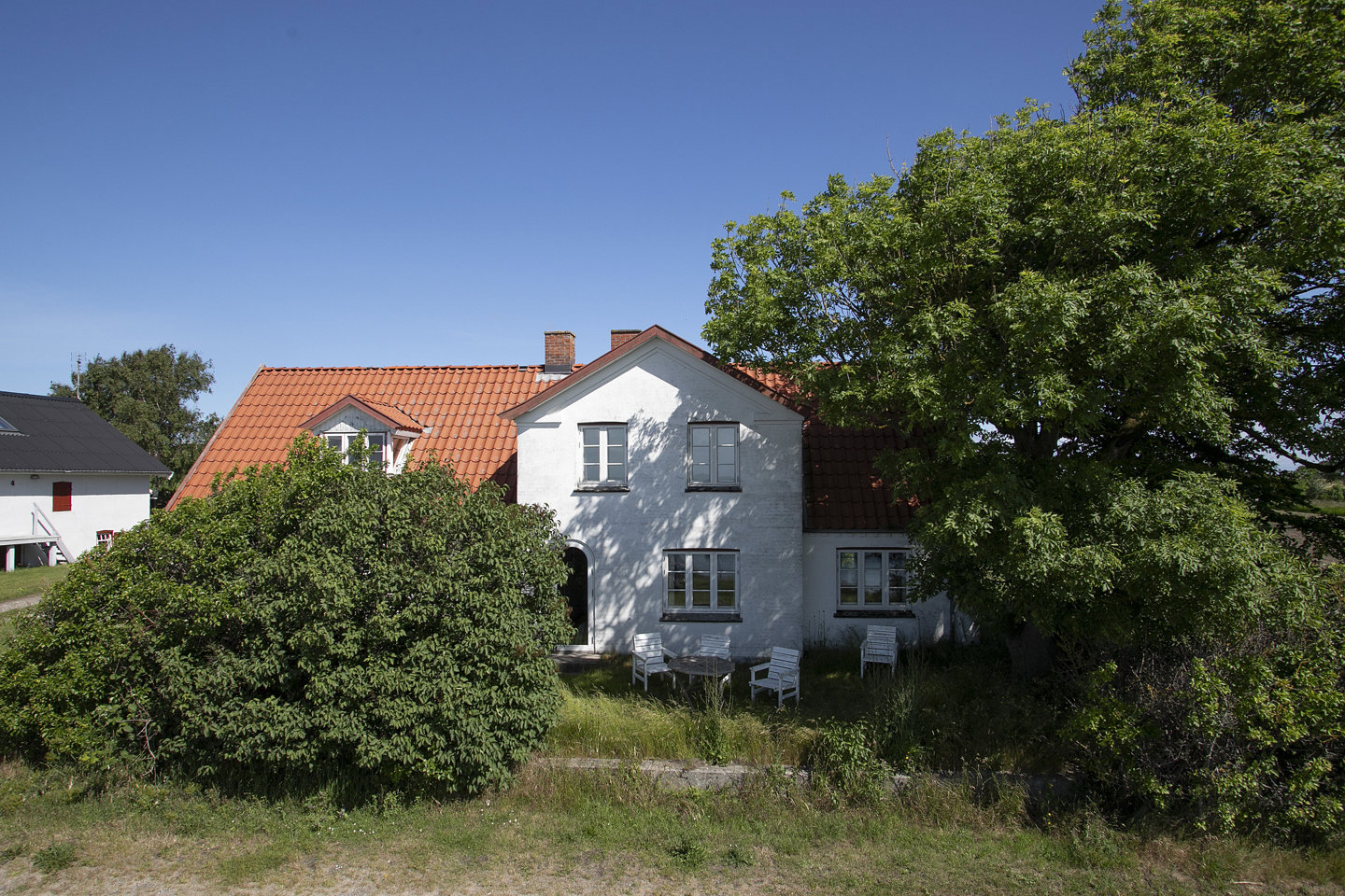 Et hvidt hus med rødt tegltag, omgivet af grønne buske og træer, står under en klar blå himmel. Tæt ved huset ses en hvid stol og et bord.