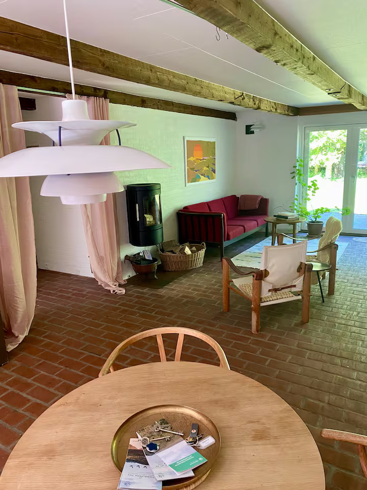 En stue med murstensgulv, træbjælker, en rød sofa, en brændeovn, planter og et rundt bord med papirer og nøgler. Sollyset strømmer ind gennem glasdørene.