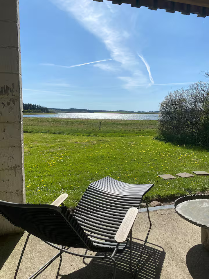 En sort lænestol og et rundt bord på en terrasse med udsigt over en græsplæne, vand og en blå himmel med tynde skyer.