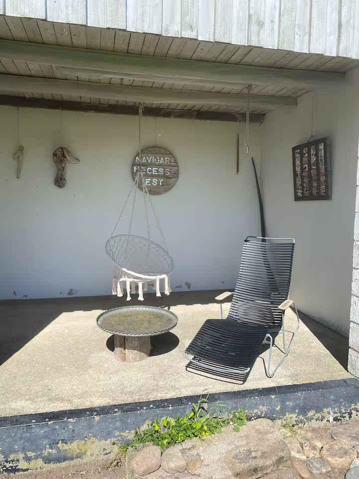 Et overdækket terrasseområde med en sort liggestol, et lille rundt bord og en hængende gyngestol. I baggrunden ses forskellige vægdekorationer.