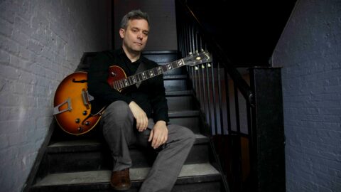 En mand i sort skjorte og grå bukser sidder på en trappe med en elektrisk guitar af mærket Sunburst i hænderne og et neutralt udtryk i ansigtet.